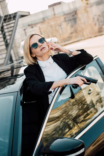 Rubia fumador en gafas de sol celebración de cigarrillo y pistola cerca de automóvil - foto de stock
