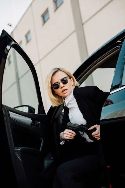 Femme blonde émotionnelle dans les lunettes de soleil tenant pistolet près de voiture noire — Photo de stock