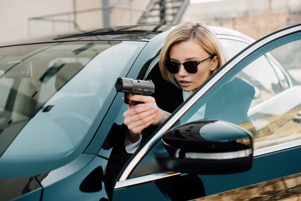 Confident blonde woman in sunglasses holding gun near black car — Stock Photo