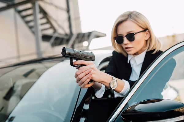 Selective focus of serious blonde woman in sunglasses holding gun near black car — Stock Photo