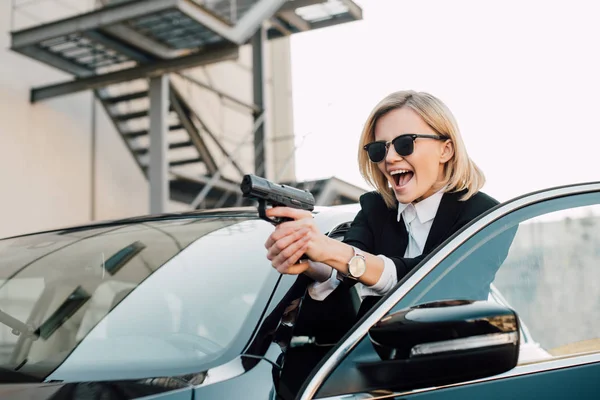 Femme blonde gaie dans les lunettes de soleil tenant pistolet près de voiture noire — Photo de stock