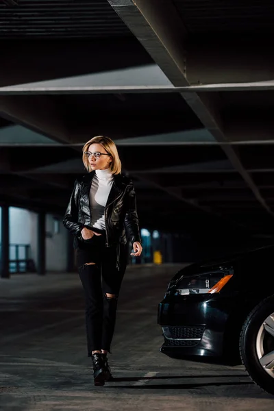 Elegante donna bionda che cammina con mano in tasca vicino auto nera nel parcheggio — Foto stock