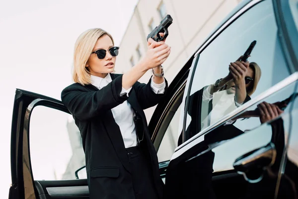 Dangerous blonde woman in sunglasses holding gun near car — Stock Photo