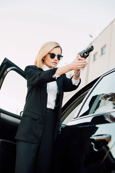 Jeune femme blonde dangereuse dans les lunettes de soleil tenant pistolet près de la voiture — Photo de stock