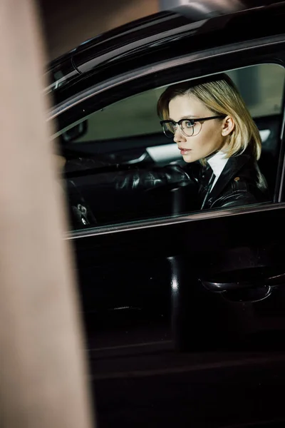 Blonde young woman in glasses sitting in black car — Stock Photo