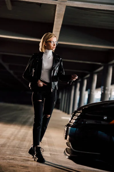 Hübsche junge blonde Frau mit Brille geht mit der Hand in der Tasche in der Nähe von Auto — Stockfoto