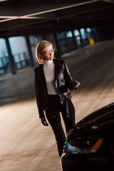 Jolie jeune femme blonde dans des lunettes tenant la clé près de la voiture — Photo de stock