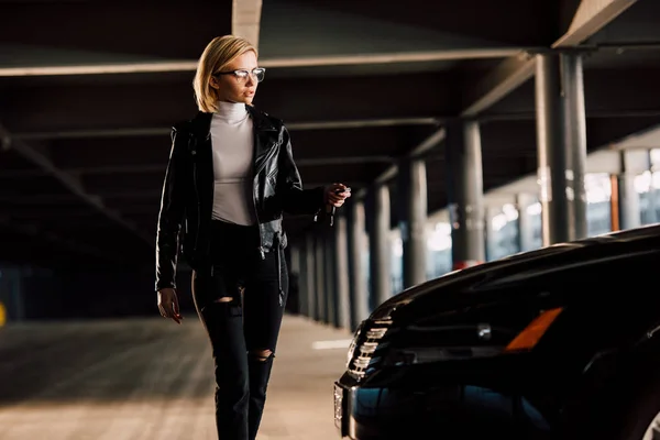 Jolie jeune femme blonde à lunettes marchant dans le parking avec clé près de la voiture — Photo de stock