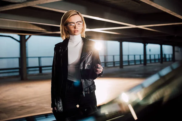 Blonde junge Frau mit Brille steht mit Autoschlüssel auf Parkplatz — Stockfoto