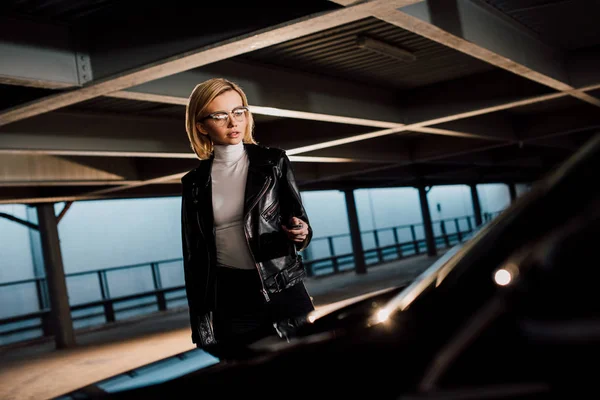 Attrayant jeune femme dans des lunettes debout dans le parking avec clé près de la voiture — Photo de stock