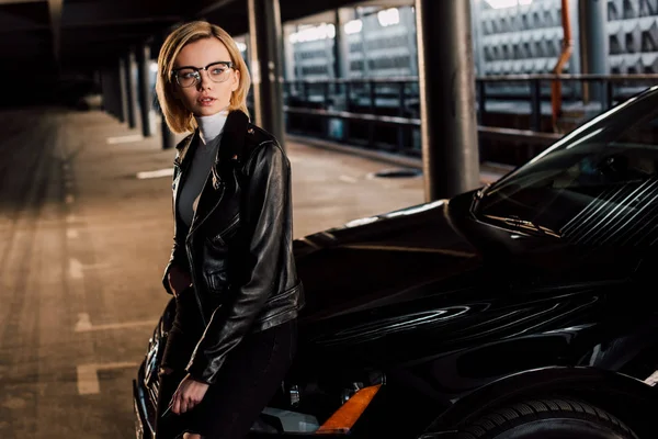 Pensosa giovane donna in occhiali in piedi nel parcheggio vicino auto nera — Stock Photo