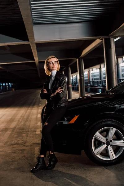 Chère jeune femme dans des lunettes debout dans le parking avec les bras croisés près de voiture noire — Photo de stock