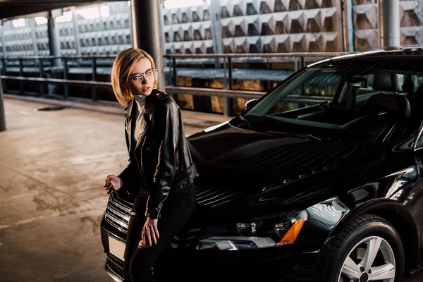 Серйозна молода жінка в окулярах стоїть на парковці біля чорної машини — стокове фото
