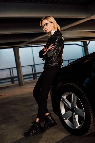 Confiant jeune femme dans des lunettes debout dans le parking avec les bras croisés près de voiture noire — Photo de stock