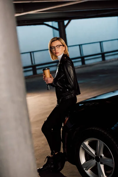 Привлекательная девушка в очках с бумажной чашкой на парковке возле черного автомобиля — стоковое фото