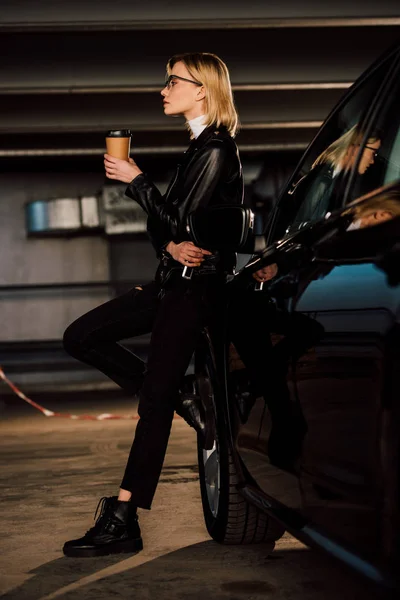 Beautiful blonde girl in glasses holding paper cup in parking near black automobile — Stock Photo