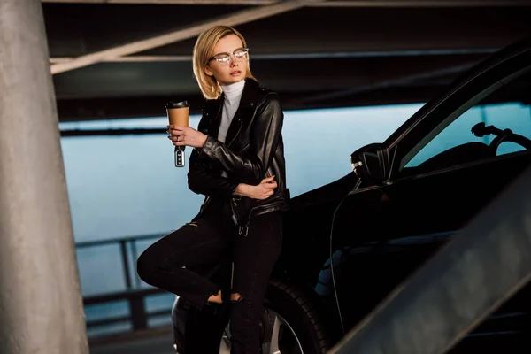 Beautiful blonde girl in glasses holding paper cup and key in parking near car — Stock Photo