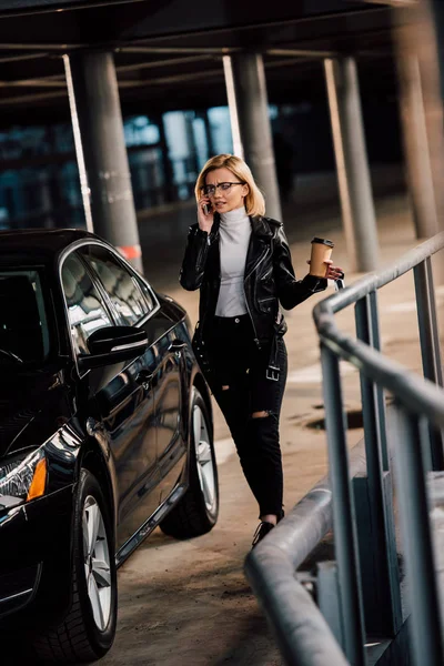 Chica rubia caminando en el aparcamiento con taza desechable y hablando en el teléfono inteligente cerca de automóvil negro - foto de stock