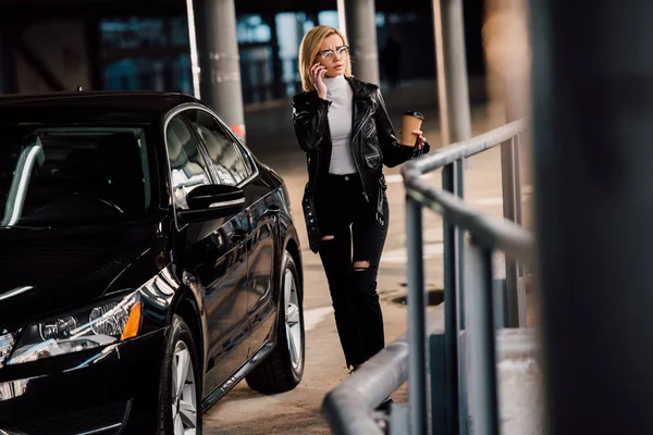 Femme blonde debout dans le parking avec tasse jetable et parler sur smartphone près de l'automobile noire — Photo de stock