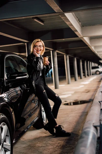 Happy blonde woman standing in parking with disposable cup while talking on smartphone near black automobile — Stock Photo