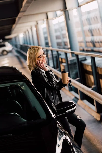 Smiling blonde woman standing in parking with disposable cup while talking on smartphone near black automobile — Stock Photo