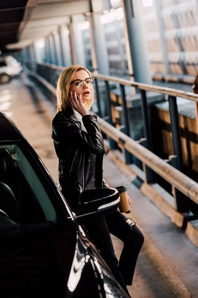 Frau beim Telefonieren in der Nähe von schwarzem Auto mit Einwegbecher auf Parkplatz überrascht — Stockfoto