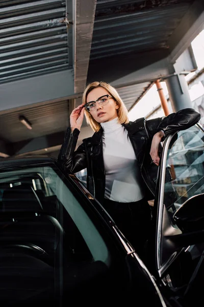 Впевнена приваблива дівчина торкається окулярів біля чорного автомобіля в парковці — стокове фото