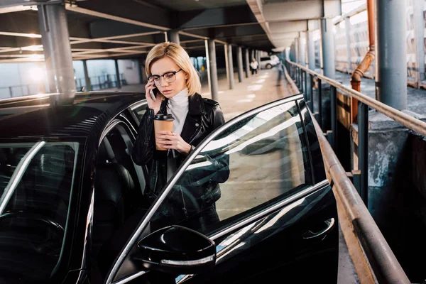 Серьезная девушка в очках разговаривает по смартфону и держит кофе, чтобы подойти к черной машине — стоковое фото