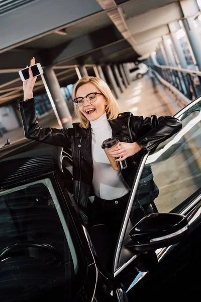 Chica feliz sosteniendo teléfono inteligente y taza desechable y agitando la mano cerca del coche - foto de stock