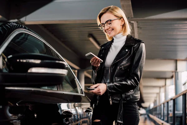 Chica alegre y atractiva sosteniendo teléfono inteligente y llave cerca de coche negro - foto de stock