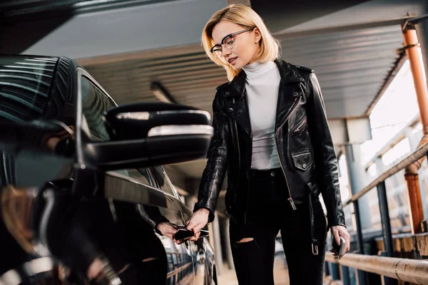 Blonde girl holding key and touching car door in parking — Stock Photo