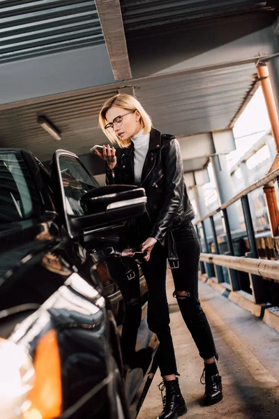 Blonde attractive girl looking at smartphone while standing near car — Stock Photo