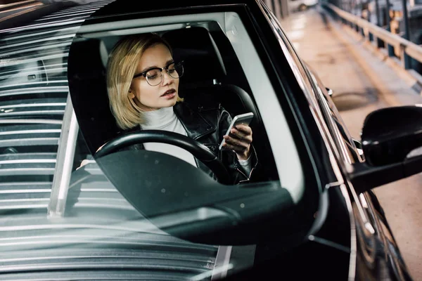 Blonde attractive woman looking at smartphone while holding steering wheel in car — Stock Photo
