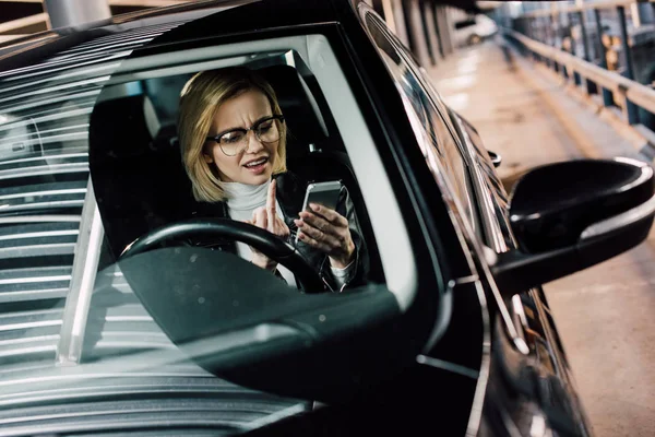 Emotional girl in glasses showing middle finger while looking at smartphone in car — Stock Photo