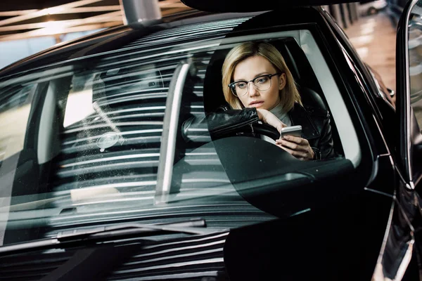 Upset blonde girl in glasses holding smartphone in car — Stock Photo