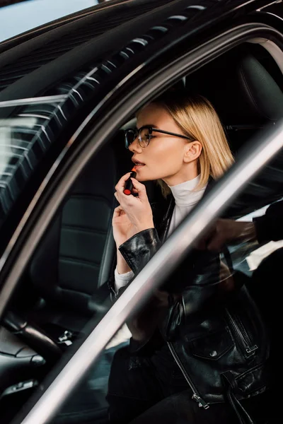 Atractiva chica rubia en gafas aplicando lápiz labial en el coche - foto de stock