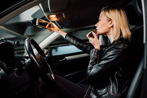 Low angle view of attractive blonde girl applying lipstick and touching mirror in car — Stock Photo