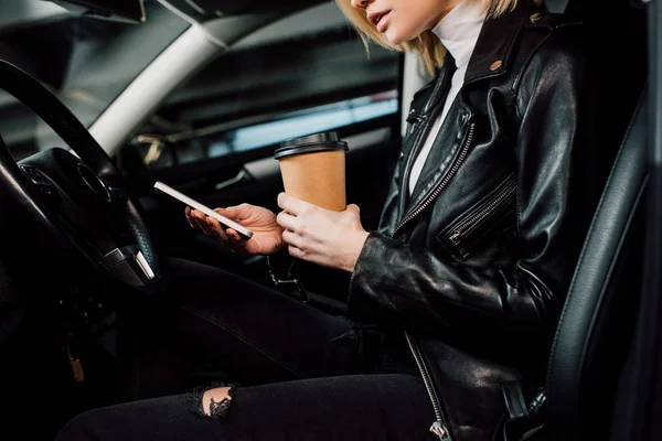 Vista recortada de niña sosteniendo taza de papel y el uso de teléfono inteligente en el automóvil - foto de stock