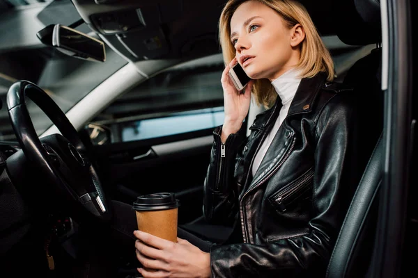 Chica rubia soñadora sosteniendo taza de papel y hablando en el teléfono inteligente en el coche - foto de stock