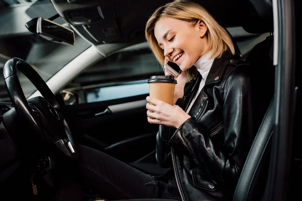 Cheerful blonde girl holding paper cup and talking on smartphone in car — Stock Photo