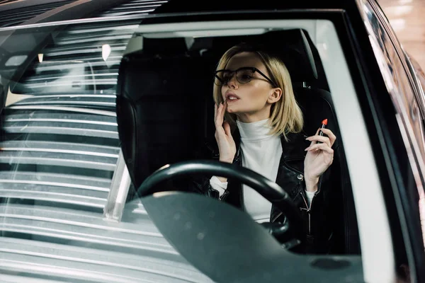 Pretty blonde girl in glasses sitting in car and applying lipstick — Stock Photo