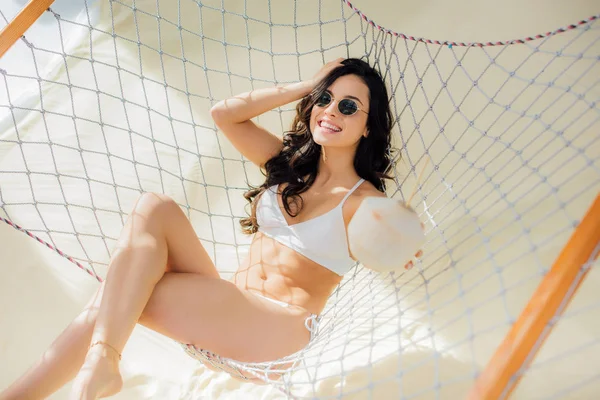 Hermosa mujer joven feliz en bikini y gafas de sol con cóctel de coco y tumbado en hamaca en la playa - foto de stock