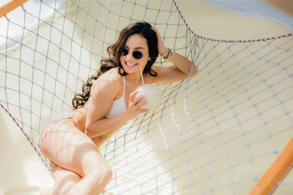 Beautiful happy girl in bikini and sunglasses holding coconut cocktail and lying in hammock on beach — Stock Photo