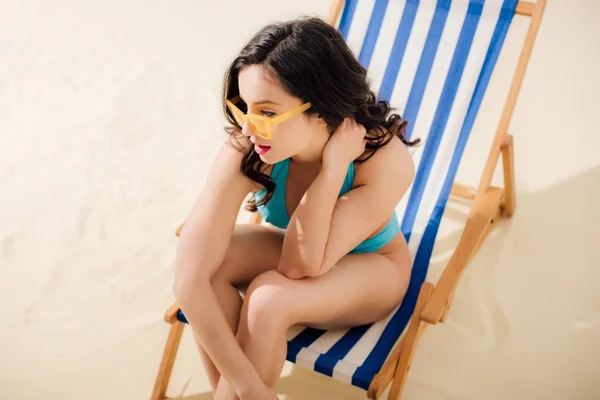 Beautiful girl in bikini and sunglasses sitting on deck chair on beach — Stock Photo