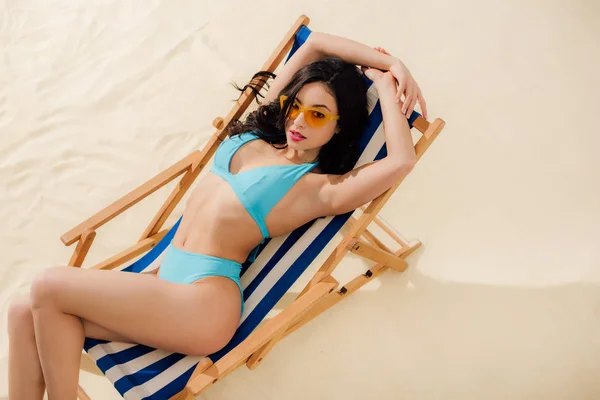 Hermosa chica en bikini y gafas de sol tumbado en la silla de cubierta y mirando a la cámara en la playa - foto de stock