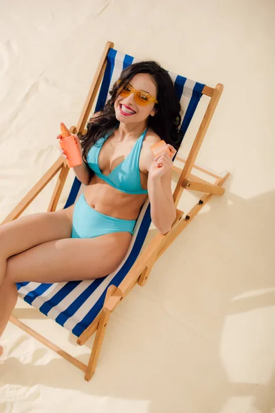 Hermosa chica sonriente en bikini sosteniendo botella de protector solar y relajarse en la silla de cubierta en la playa - foto de stock