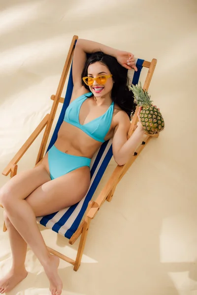 Beautiful happy girl in sunglasses and bikini holding pineapple and relaxing on deck chair on beach — Stock Photo