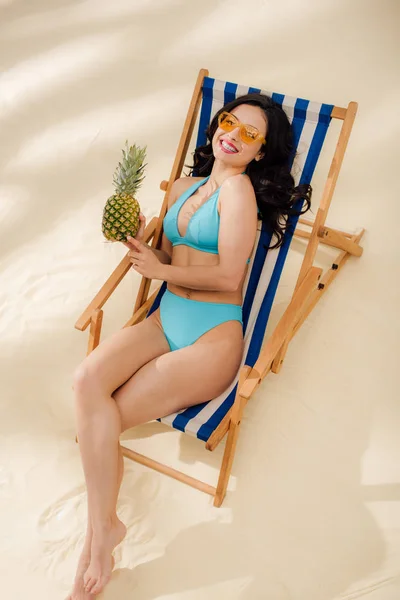 Belle fille souriante en lunettes de soleil et bikini tenant l'ananas et relaxant sur chaise longue sur la plage — Photo de stock