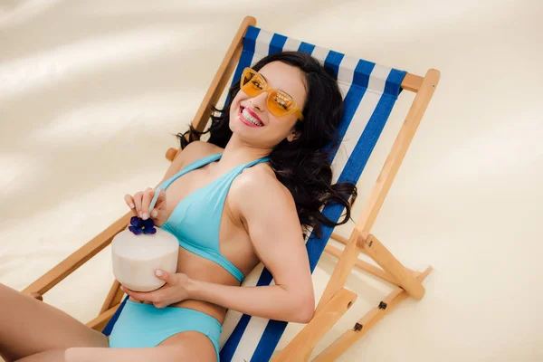 Bela menina feliz em biquíni e óculos de sol com coquetel de coco deitado na cadeira deck na praia — Fotografia de Stock