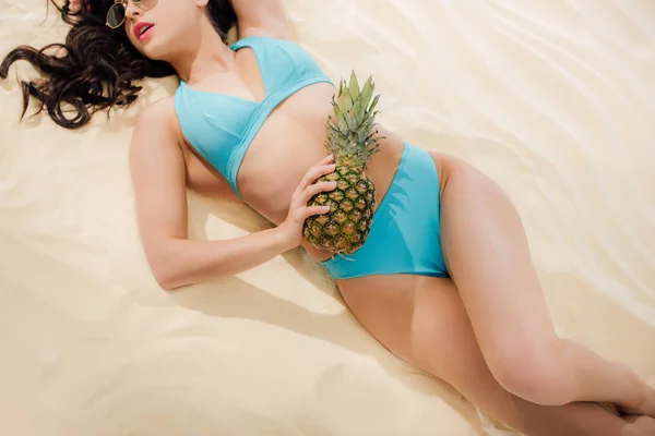 Partial view of sexy girl in blue Bikini posing with pineapple while lying on sandy beach — Stock Photo